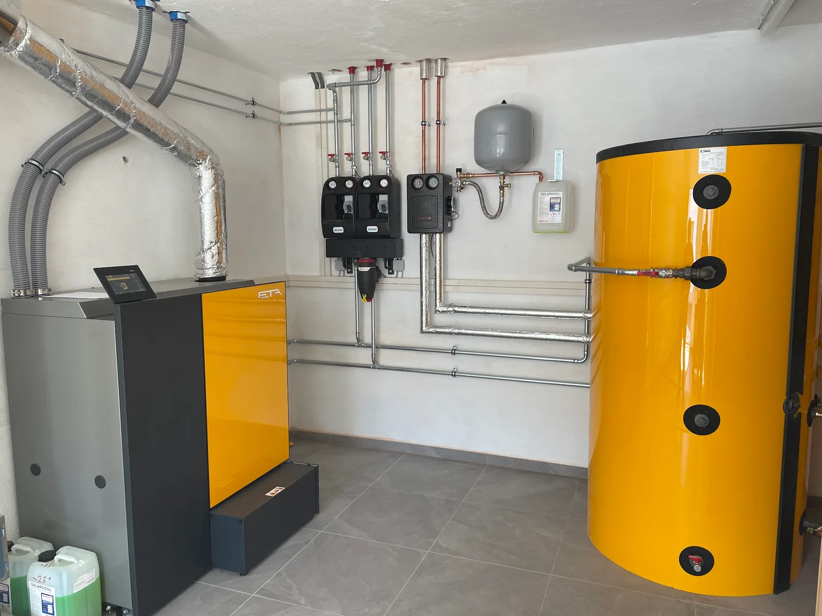 Heizraum mit gelbem Pelletskessel, Pufferspeicher und zwei Heizkreisstationen an der Wand