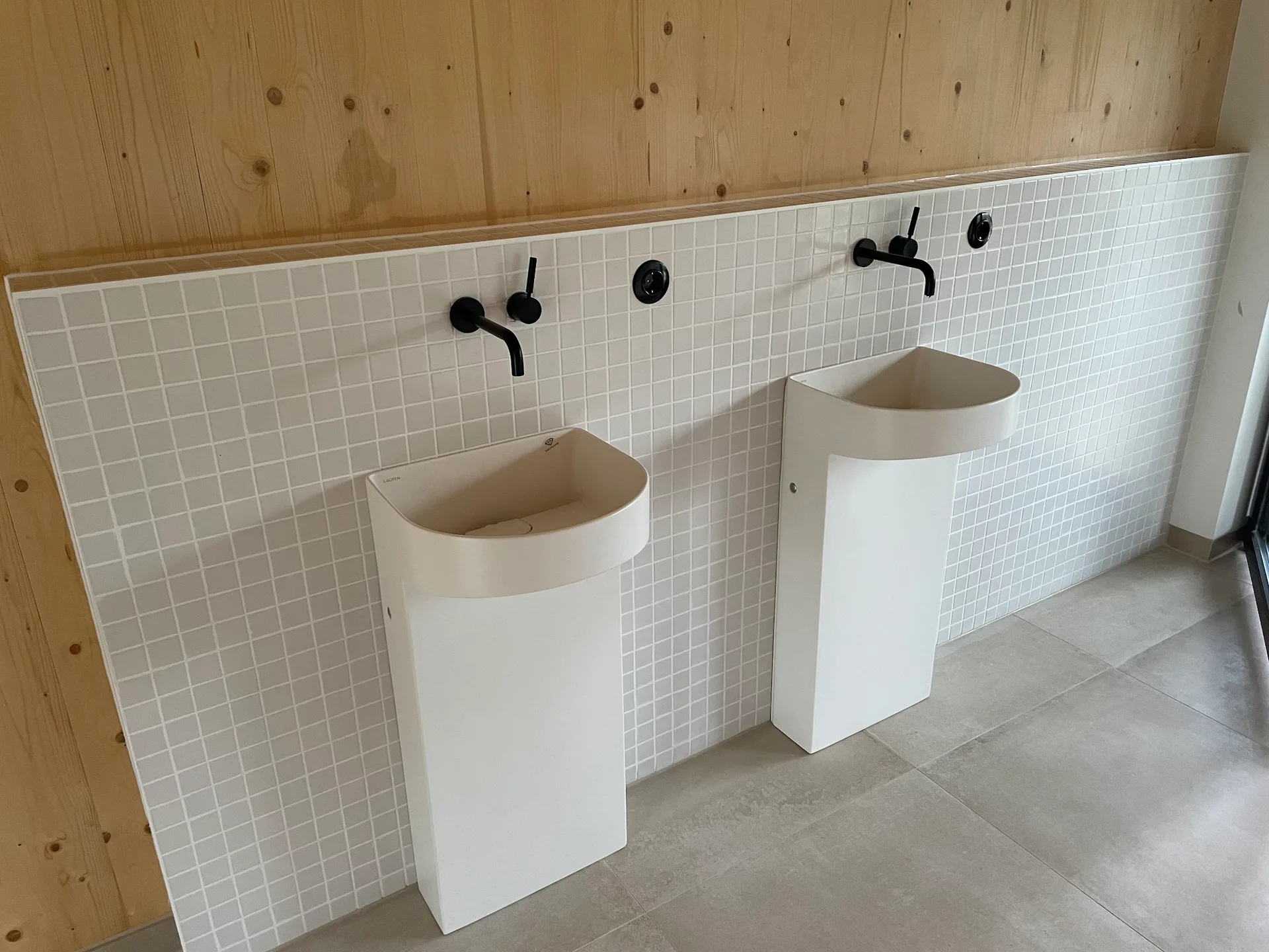 Fertiges Badezimmer mit zwei weißen Standwaschbecken, schwarzen Wandarmaturen und weißem Mosaik vor einer Holzwand
